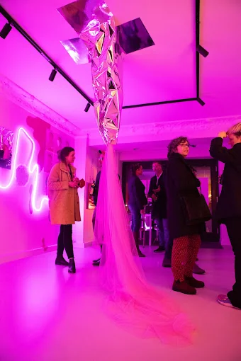 Vernissage in pinkem Licht, in der Mitte hängt eine Skulptur aus Spiegelfolie mit langem rosa Tüllschleier