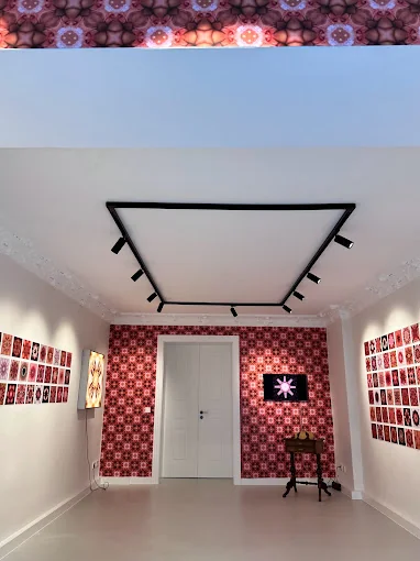 Raum mit rot-weiß-schwarzer Kaleidoskop-Tapete an der Stirnwand, an den Seitenwänden Raster kleiner Bilder im selben Muster