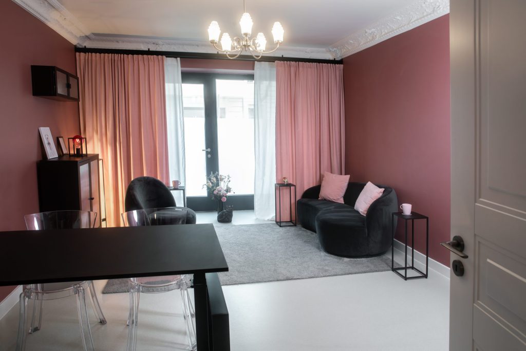 Sprechzimmer mit altrosa Wänden, schwarzem Sofa, Schreibtisch mit transparenten Stühlen und Kronleuchter