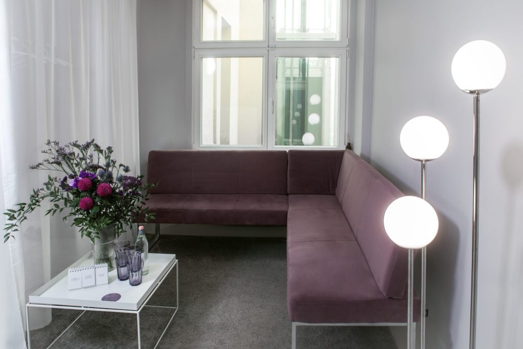 Heller Wartebereich mit violetter Eckbank, Beistelltisch mit Blumenstrauß und Wasserglas sowie drei Stehlampen mit Kugelschirmen
