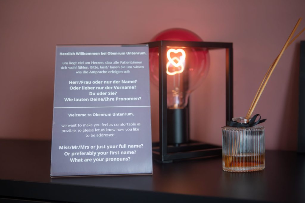 Aufsteller mit zweisprachigem Willkommenstext, der nach bevorzugter Anrede und Pronomen fragt, daneben eine Lampe mit herzförmiger Glühbirne und ein Raumduft